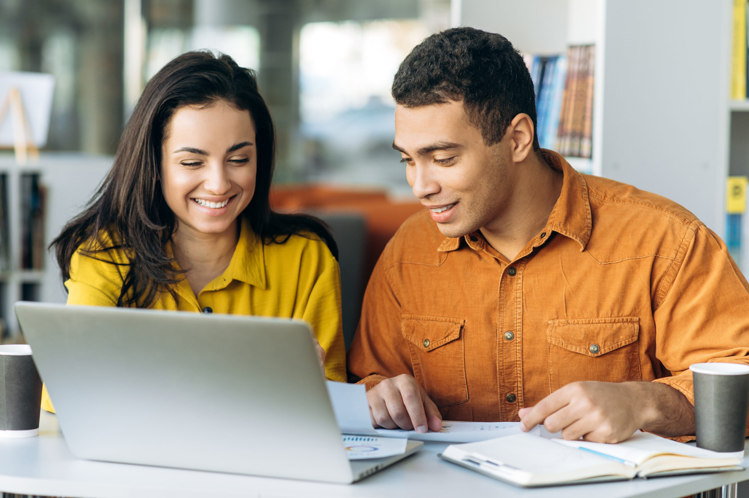 Two colleagues or students, caucasian girl and hispanic guy, are using a laptop while sitting at workplace discussing about job or education and smiling